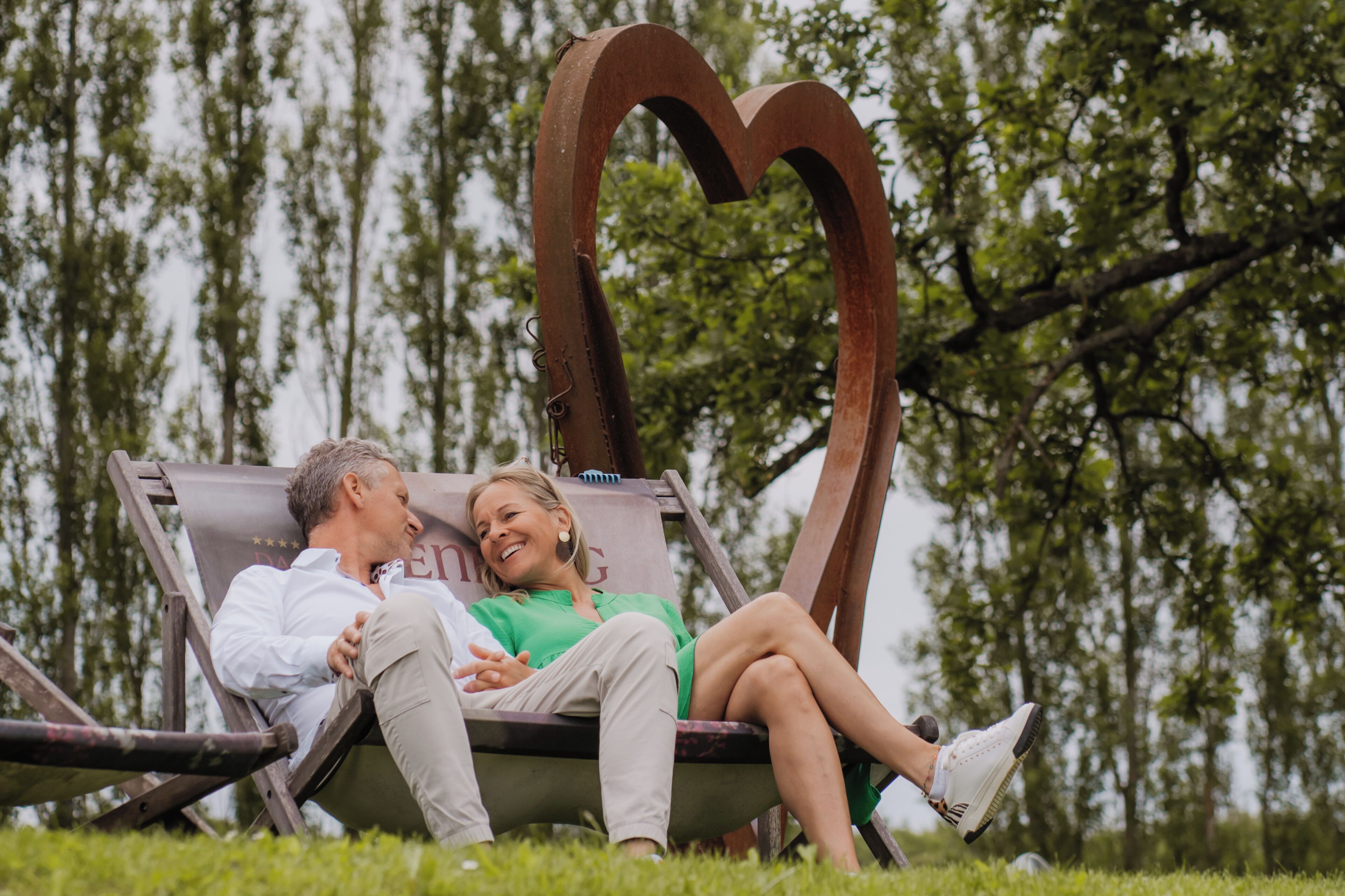 A couple sitting on a wooden bench with a heart-shaped metal sculpture behind them, in a park with trees in the background.
