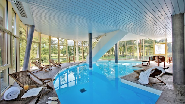 Piscina interior com paredes de vidro transparente, espreguiçadeiras de madeira e vista para o exterior.