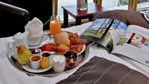 Uma bandeja de café da manhã com diversos alimentos e um copo de suco de laranja sobre uma mesa perto de uma janela com vista para um lago.