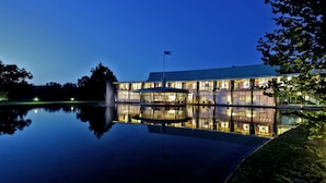 Um edifício moderno com fachada de vidro refletida em águas calmas, uma bandeira no topo e uma entrada bem iluminada.