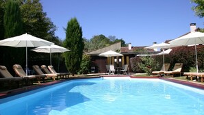 A swimming pool with lounge chairs and umbrellas, surrounded by greenery and a building.