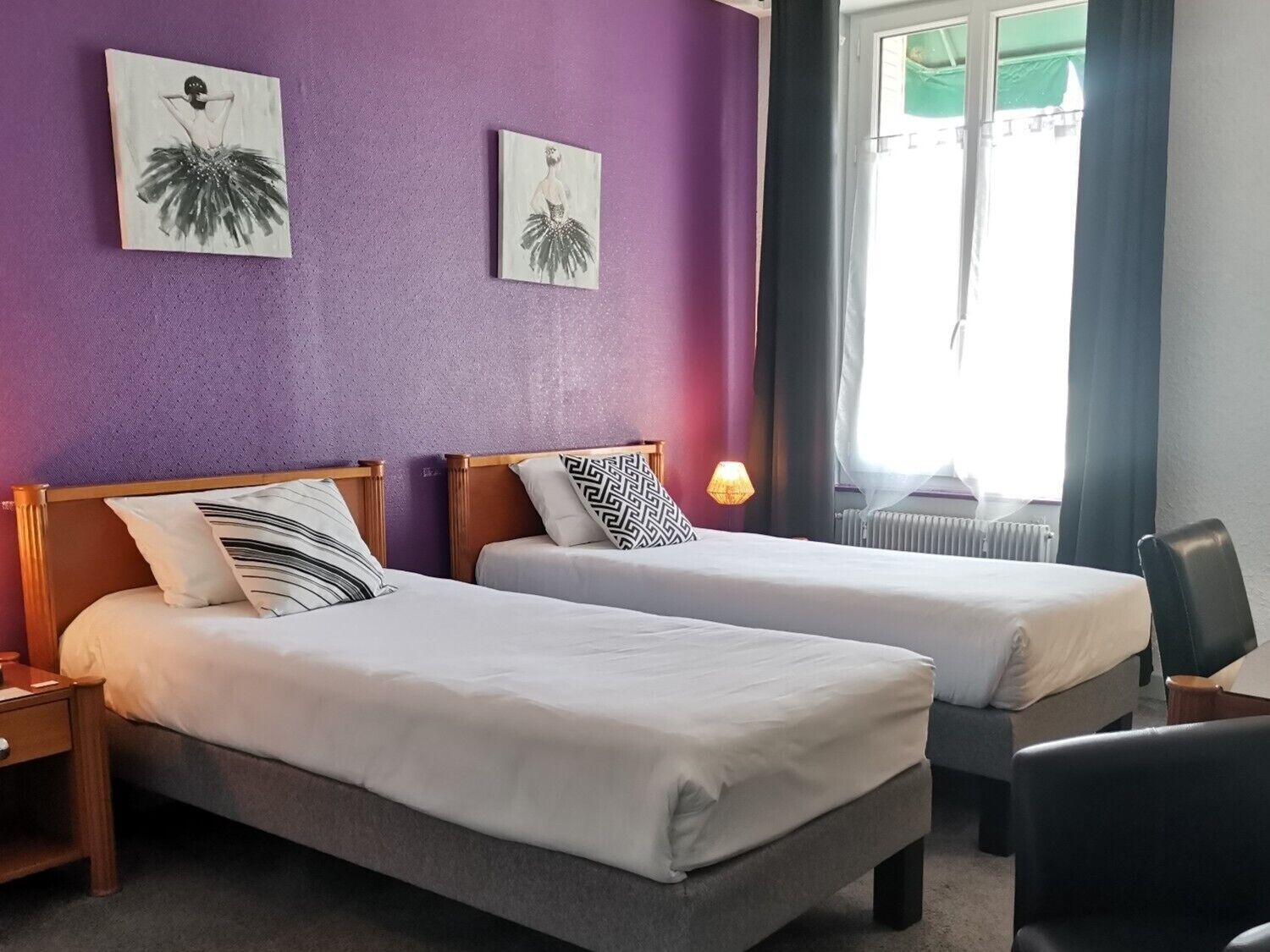 A hotel room with two single beds, a wooden headboard, a nightstand, a lamp, a chair, and two wall-mounted artworks.
