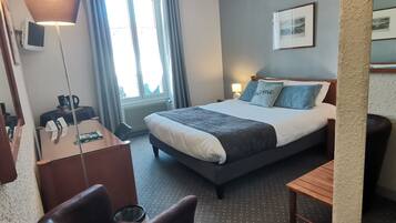 A hotel room with a bed, a desk, a chair, a lamp, and a window with curtains.
