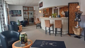 Una zona de bar con mostrador de madera, taburetes y una lámpara decorativa. En primer plano, sillas y una mesa con superficie de vidrio.