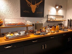 A breakfast bar with various food items, a coffee machine, and a sign indicating breakfast hours. - Hôtel Le Cerf (Briare)