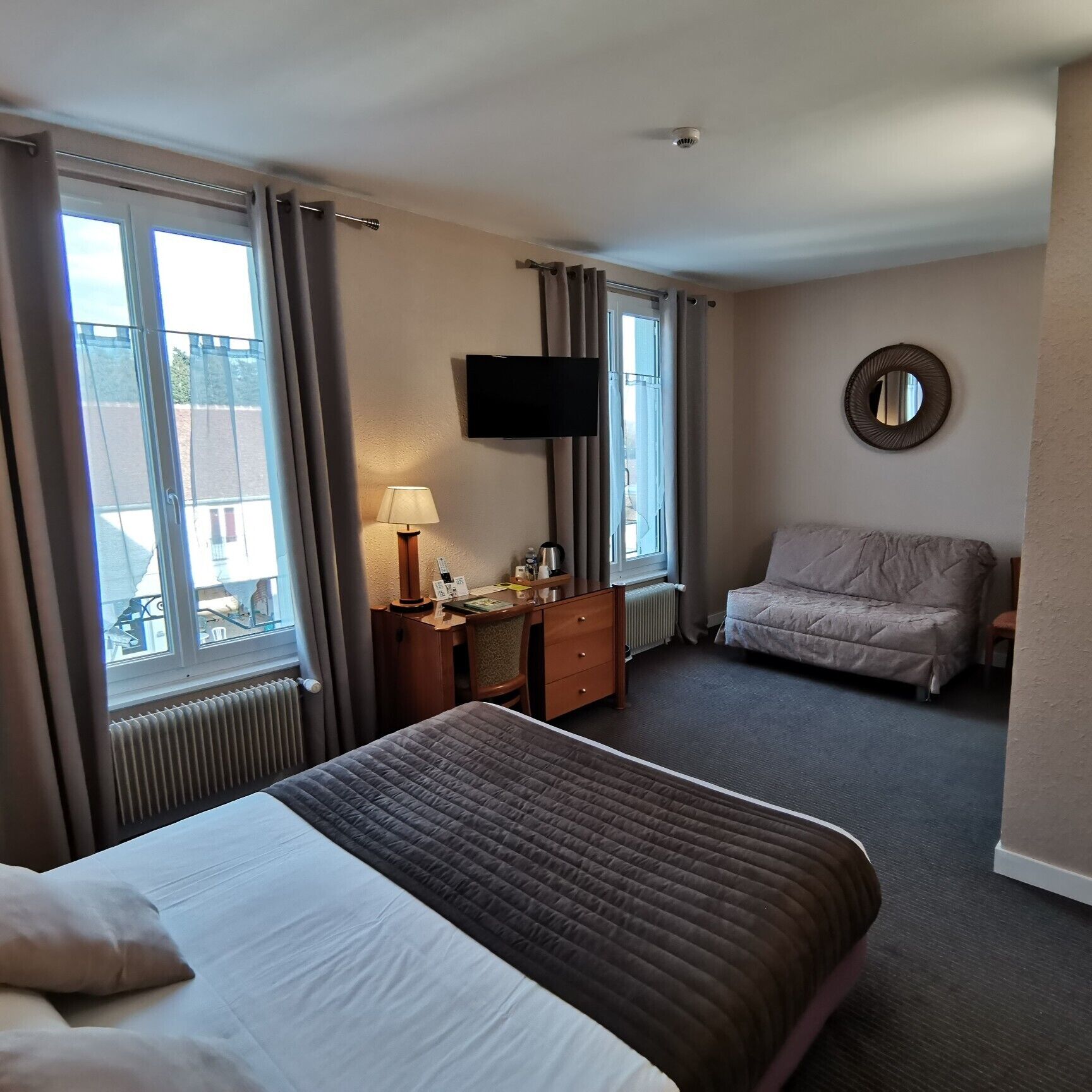A hotel room with a bed, a desk with a lamp, a sofa, a TV, and a window with curtains.