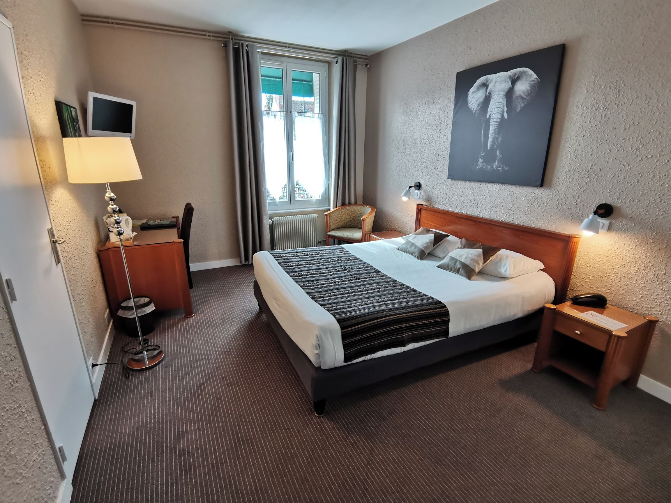 Een hotelkamer met een groot bed, een bureau met lamp, een stoel, een raam met gordijnen, een radiator en een aan de muur hangend beeld van een olifant.