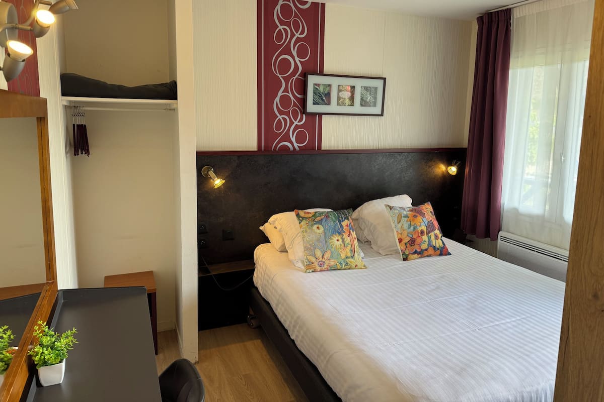 A hotel room with a bed, bedside table, and a small desk with a chair. There is a potted plant on the desk.