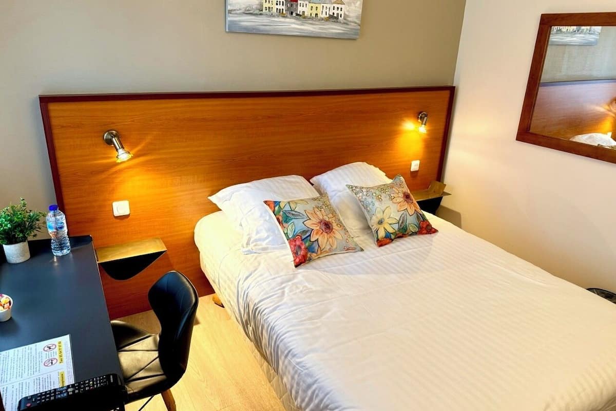 A hotel room with a bed, a desk, a chair, a water bottle, and a painting on the wall.