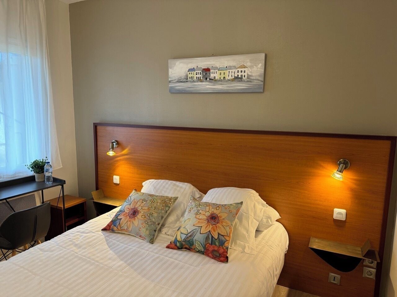 A hotel room with a bed, two pillows, a nightstand, a lamp, a painting on the wall, and a window with curtains.
