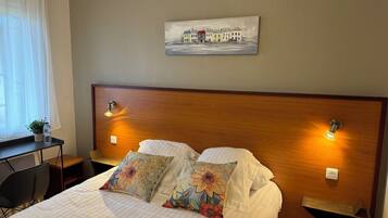 A hotel room with a bed, two pillows, a nightstand, a lamp, a painting on the wall, and a window with curtains.