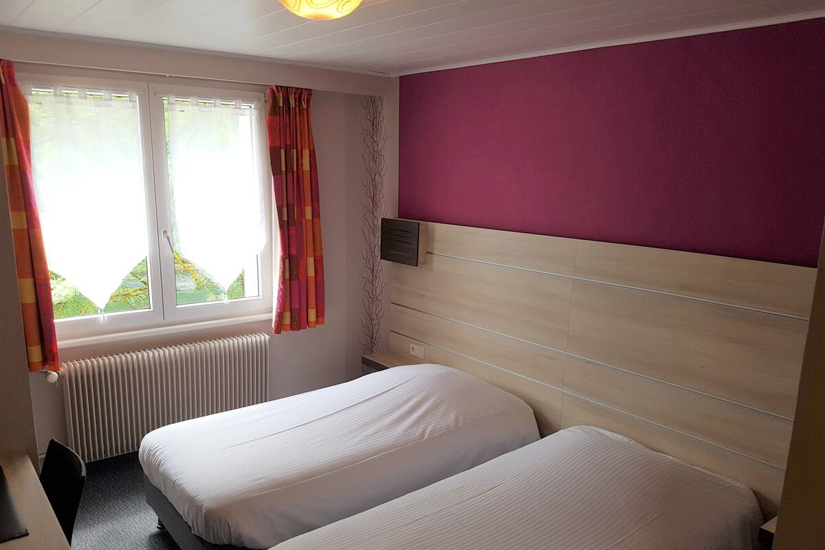 Ein Hotelzimmer mit zwei Einzelbetten, einem Fenster mit Vorhängen, einem Heizkörper und einer Wand mit Holzpaneelen.