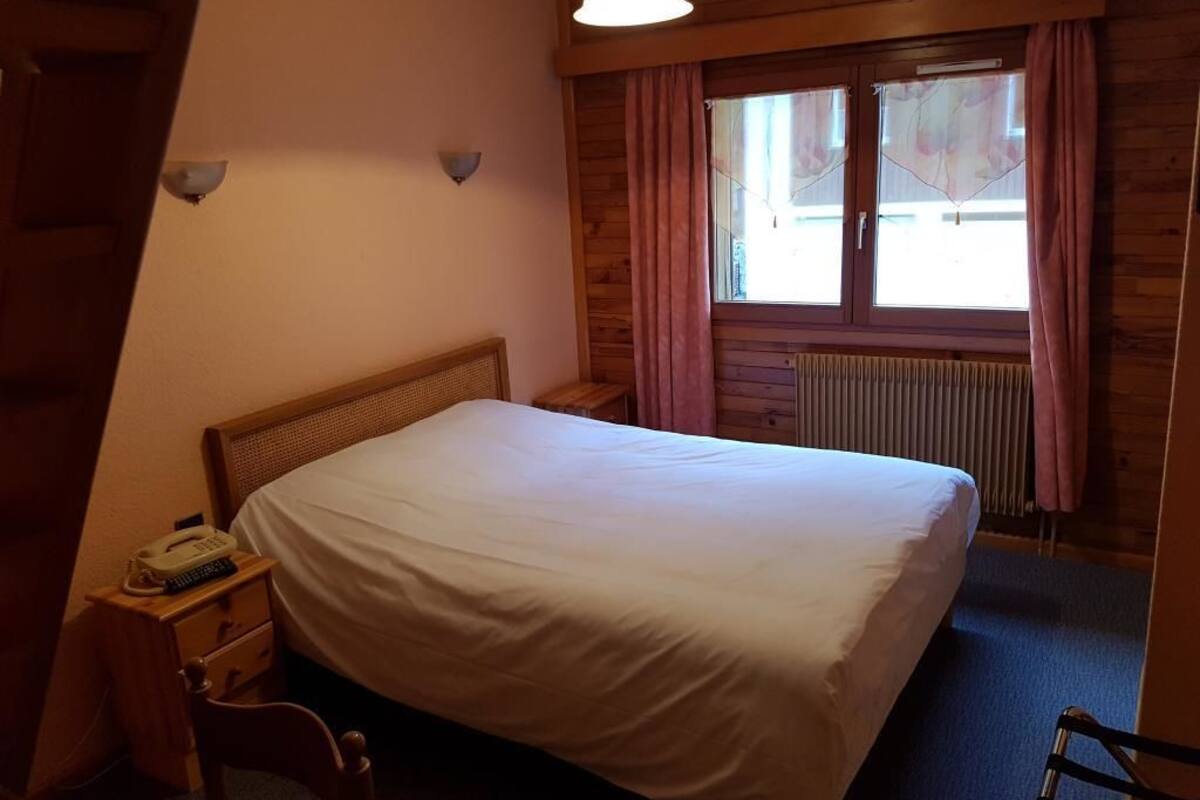 Ein Schlafzimmer mit einem Bett, einem Nachttisch, einem Sessel, einem Fenster mit Vorhängen und einer Holzbalkendecke.