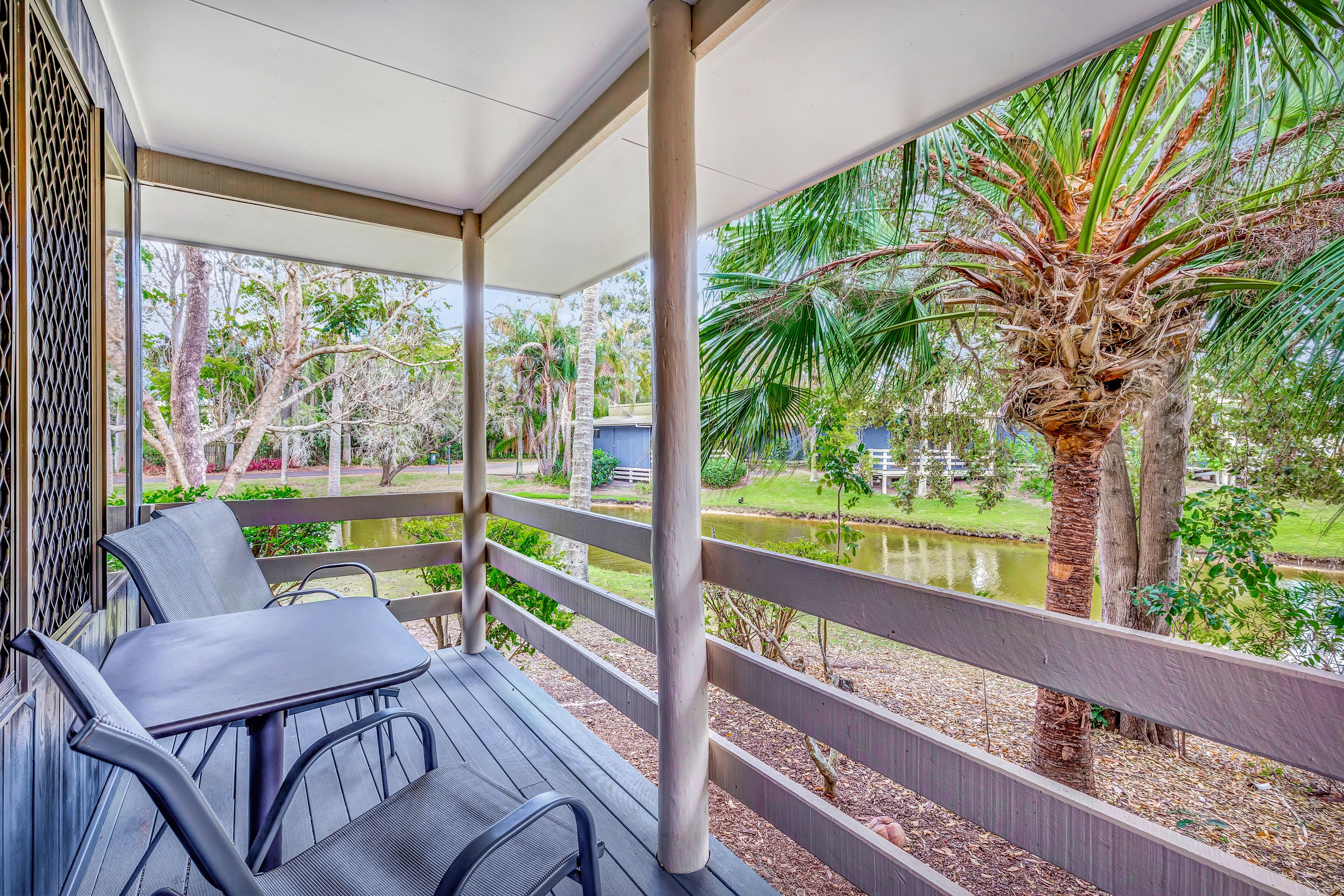 A covered outdoor seating area with a table and chairs, overlooking a pond and palm trees.