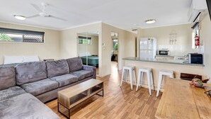 A modern living room with a grey sofa, wooden coffee table, and a kitchen area with a fridge, microwave, and bar stools. - Sanctuary Lakes Fauna Retreat (Urangan)