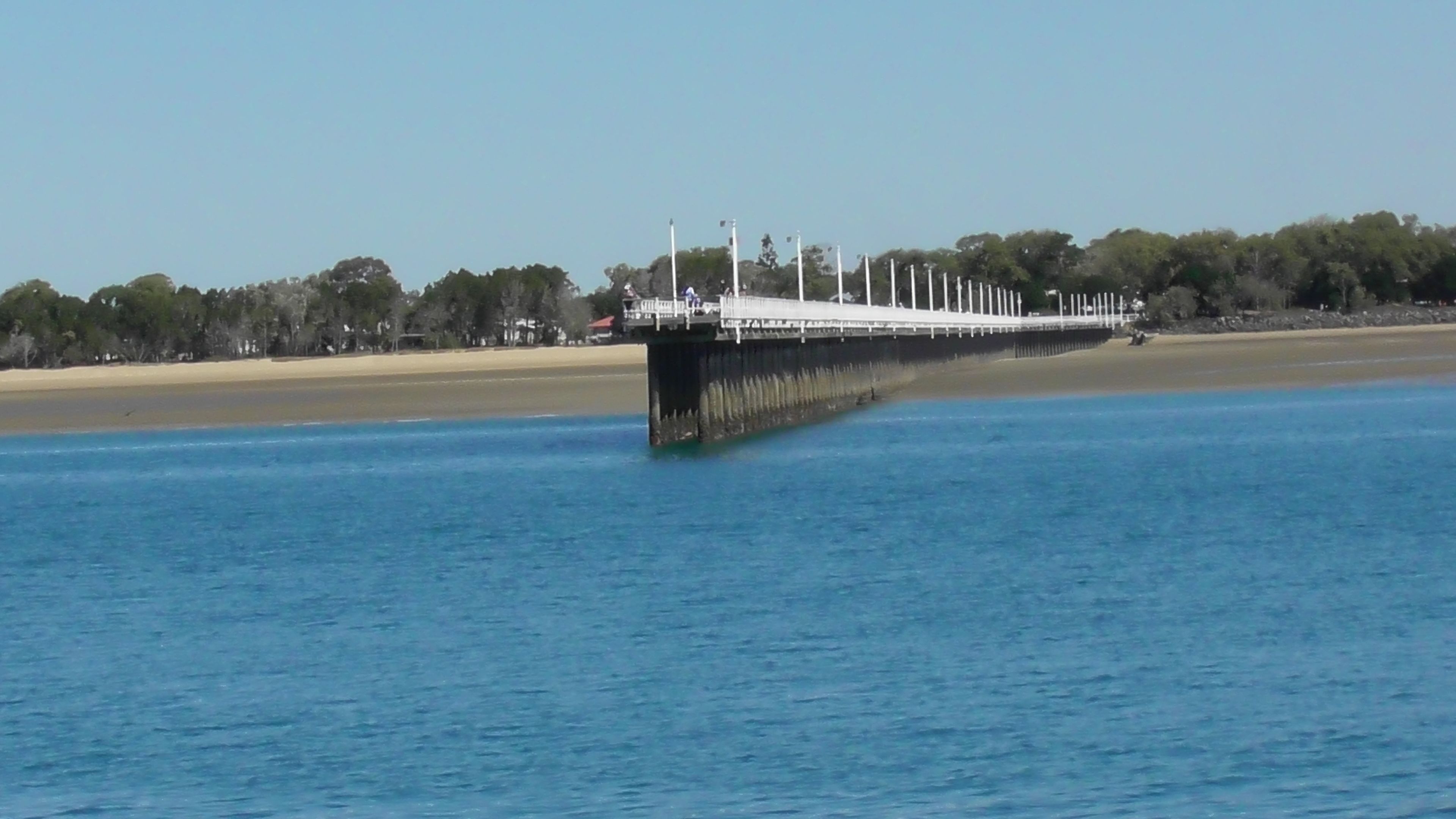 Un barco grande atracado en un muelle, con una playa arenosa y árboles al fondo.
