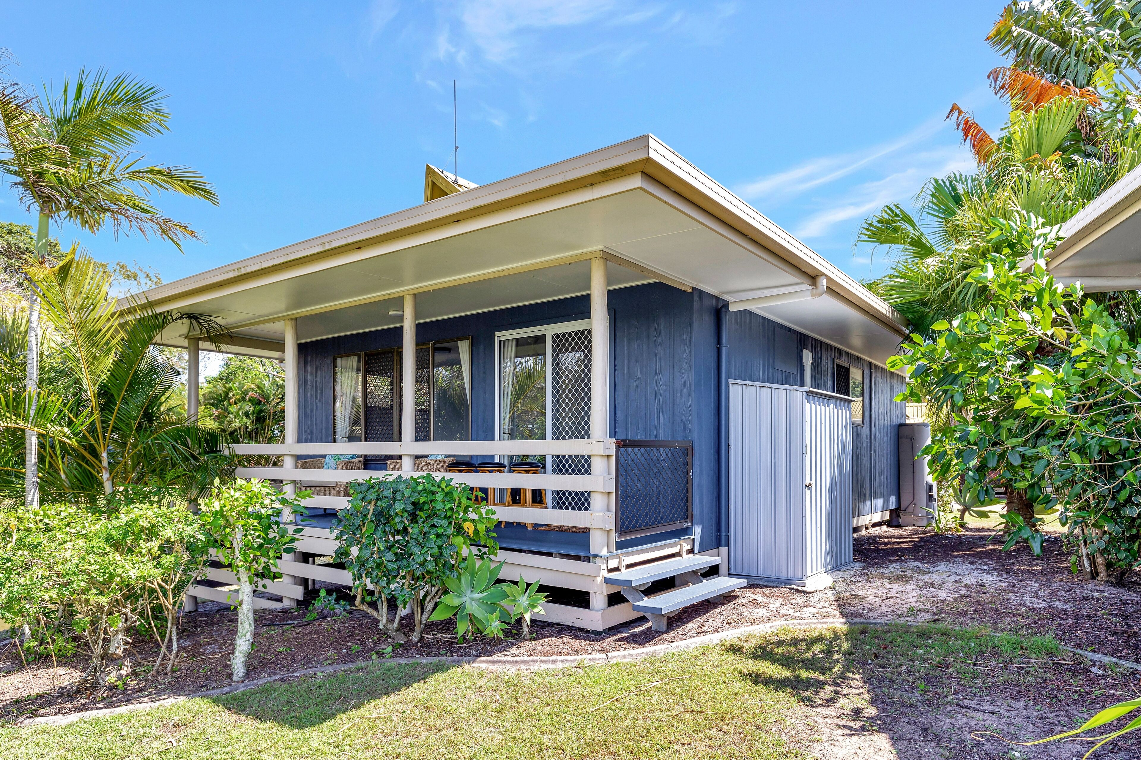 A single-story house with a covered porch, surrounded by lush greenery and palm trees.