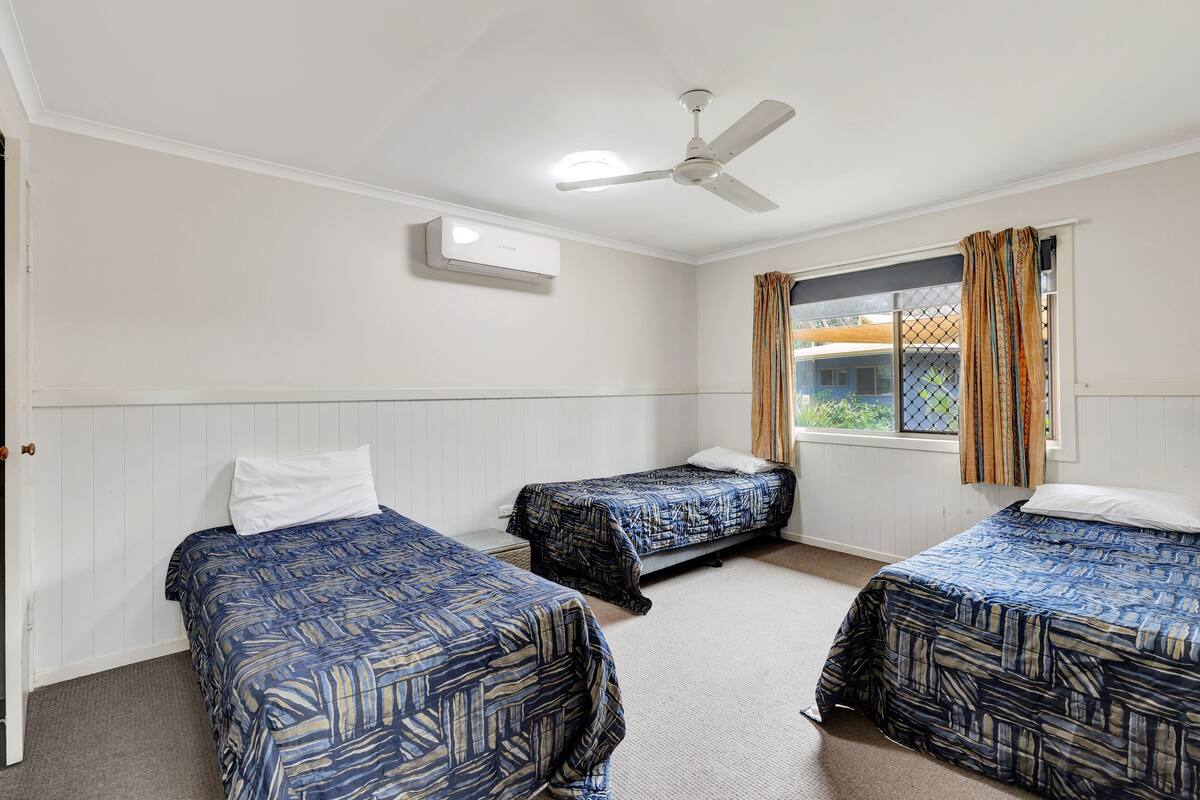 Una habitación con tres camas, ventilador de techo, aire acondicionado y una ventana con cortinas.