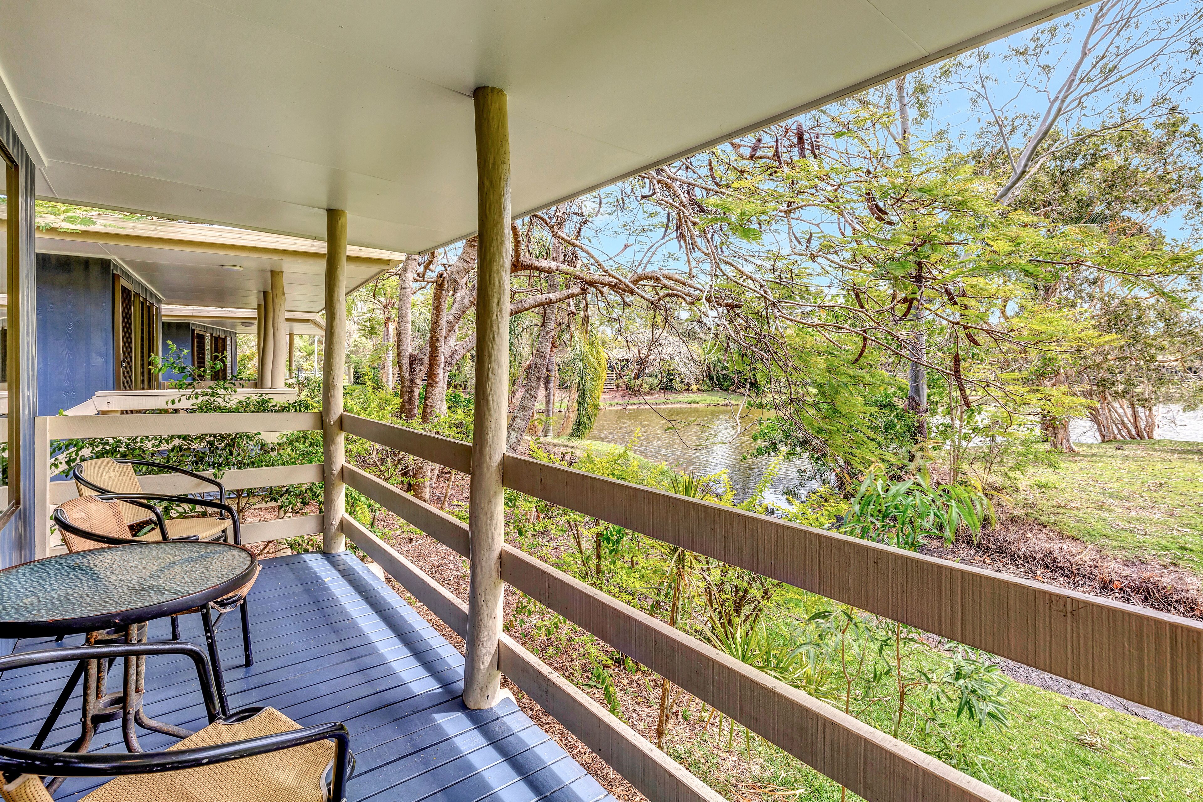 A covered outdoor deck with a table and chairs, overlooking a pond and greenery.