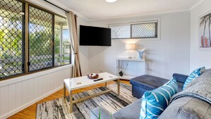 A living room with a white table, a grey sofa, a TV mounted on the wall, and a window with a view of palm trees. - Sanctuary Lakes Fauna Retreat (Urangan)