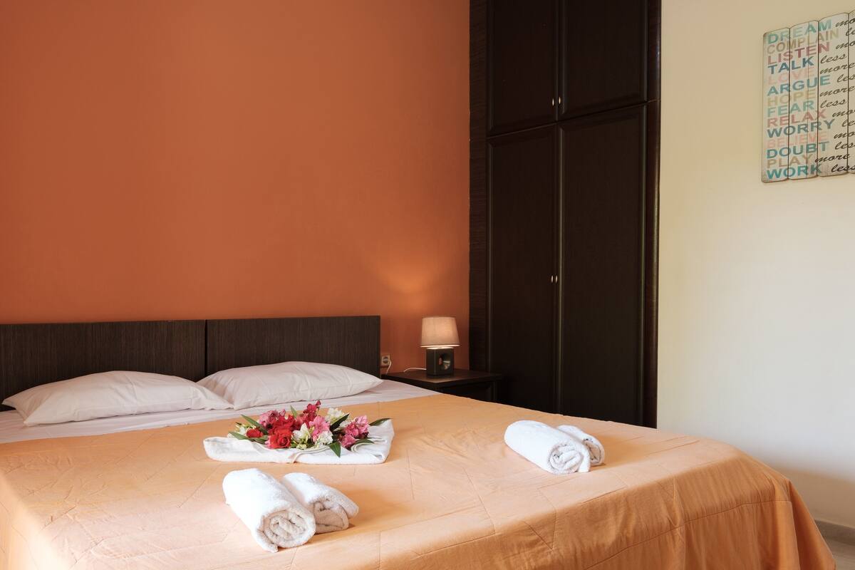 A hotel room with a bed, pillows, a nightstand with a lamp, a bouquet of flowers, and a wall with motivational words.