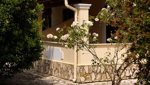 A building with a stone base and a cream-colored upper section, featuring a red roof and a white column. - Erifili Apartments (Corfu)