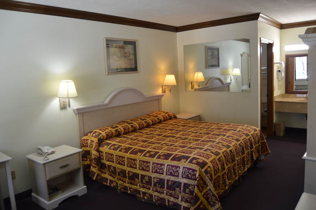A hotel room with a bed, bedside table, lamp, mirror, and a bathroom visible through an open door.