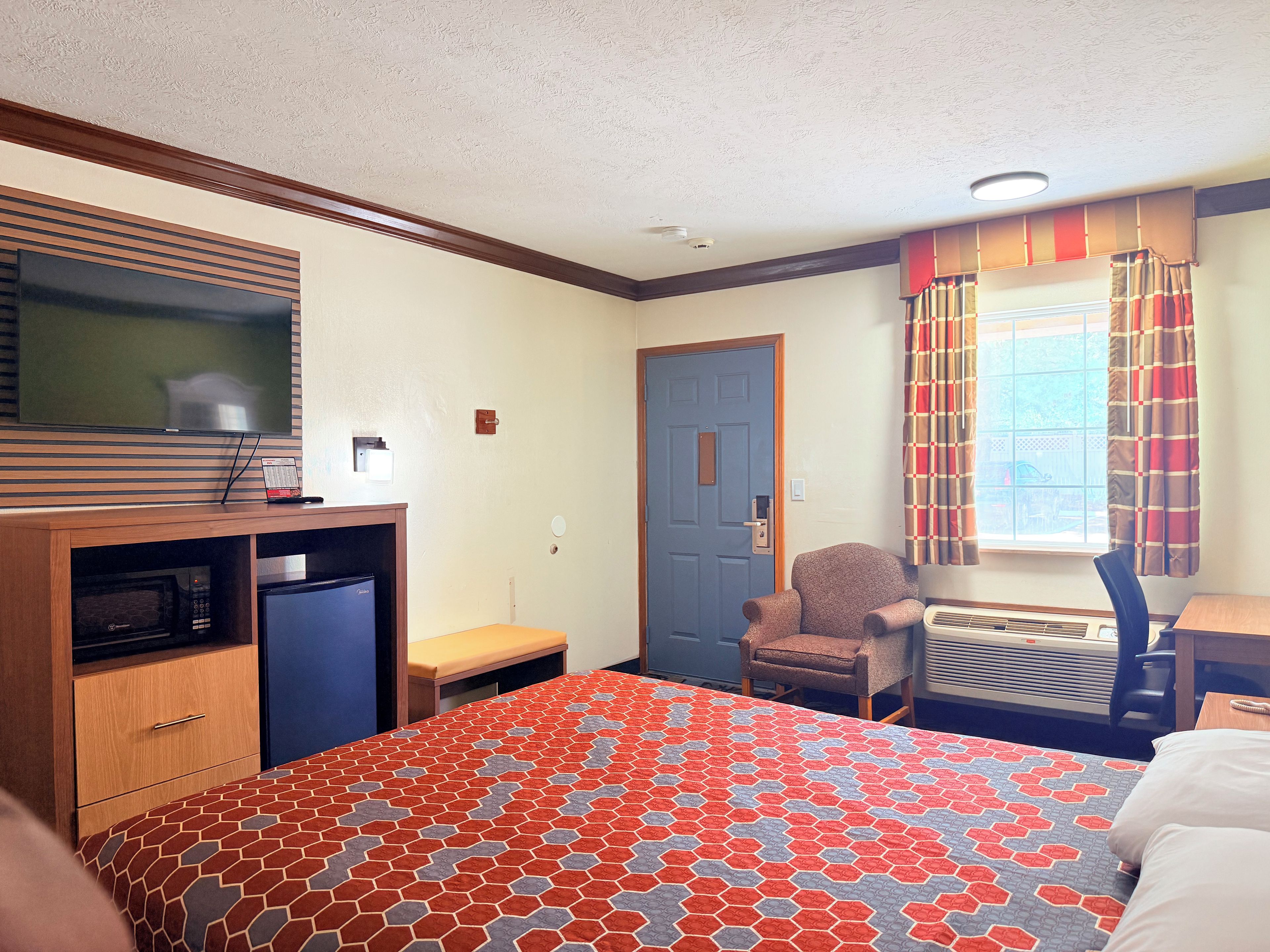 Ein Hotelzimmer mit einem Bett, einem Schreibtisch, einem Stuhl, einem Fernseher und einem fensterbepflanzten Fenster.
