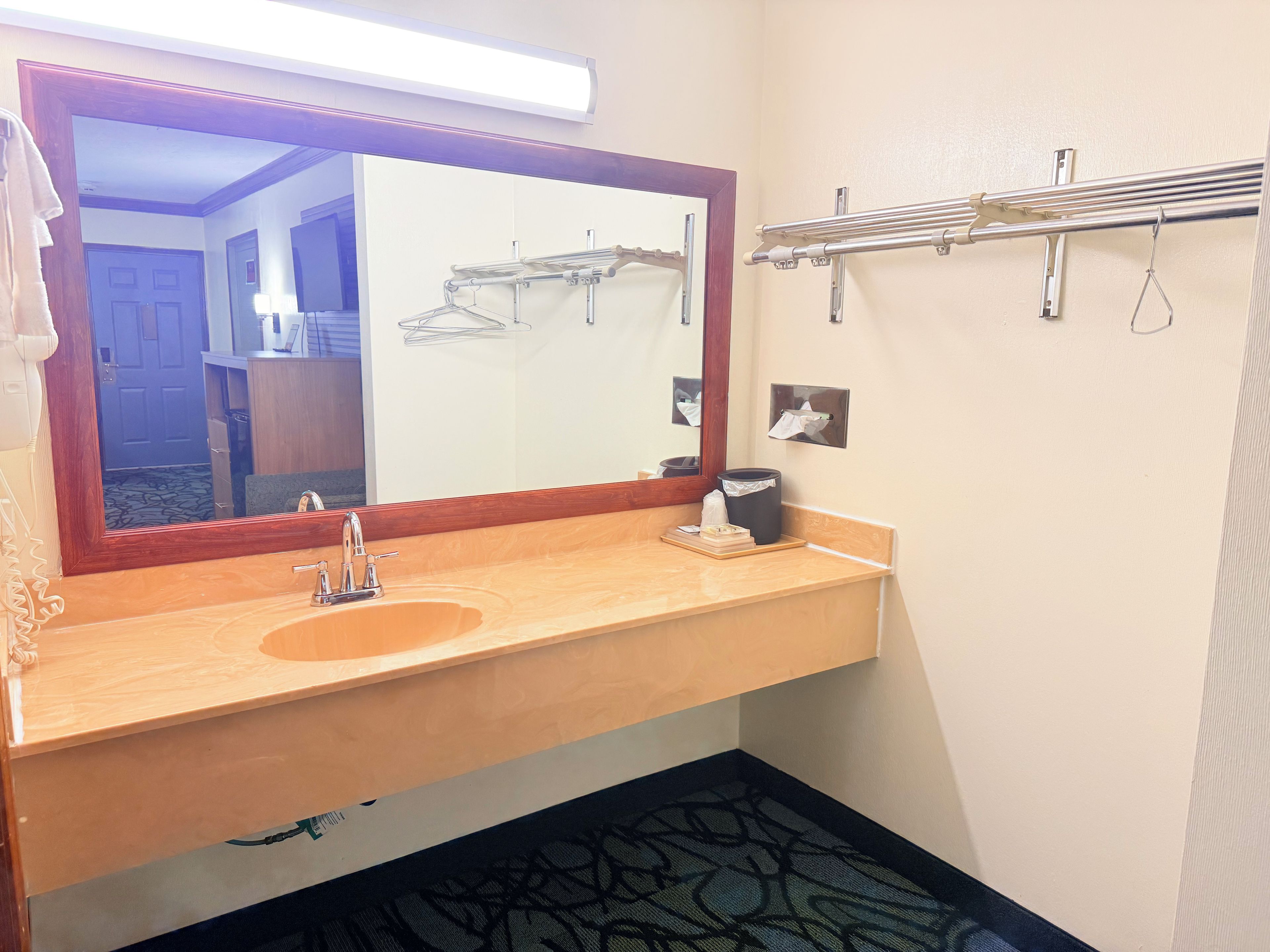 A bathroom with a wooden vanity, a large mirror, a towel rack, and a trash bin.
