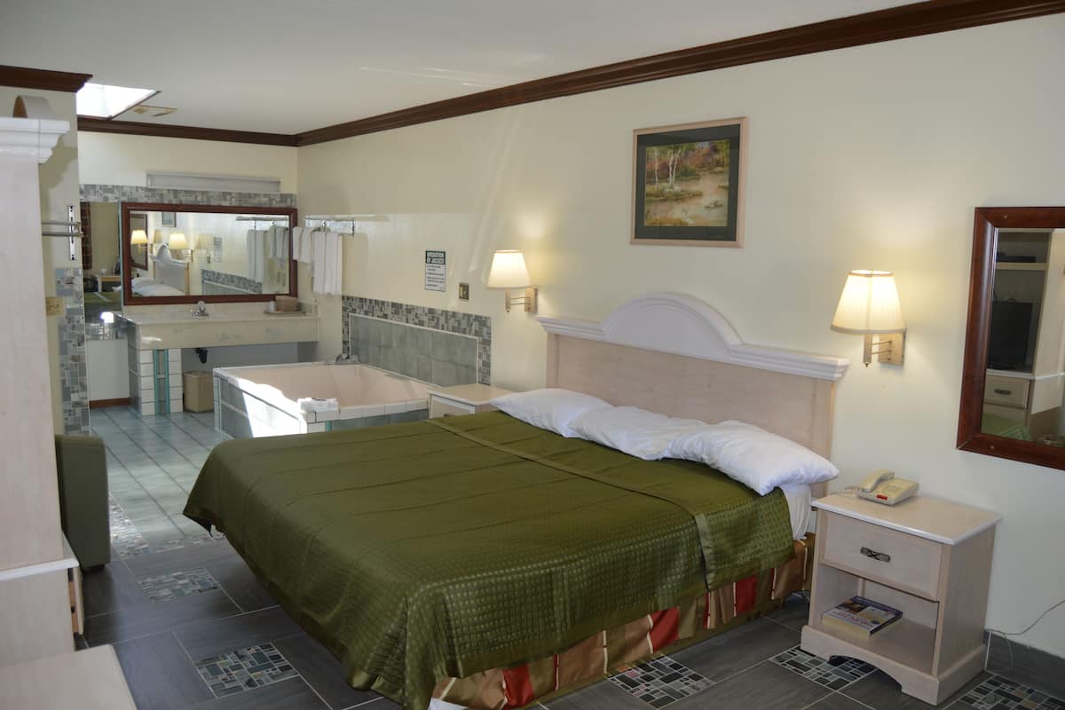 A hotel room with a bed, bedside table, nightstand, lamp, and a bathroom with a sink and mirror.