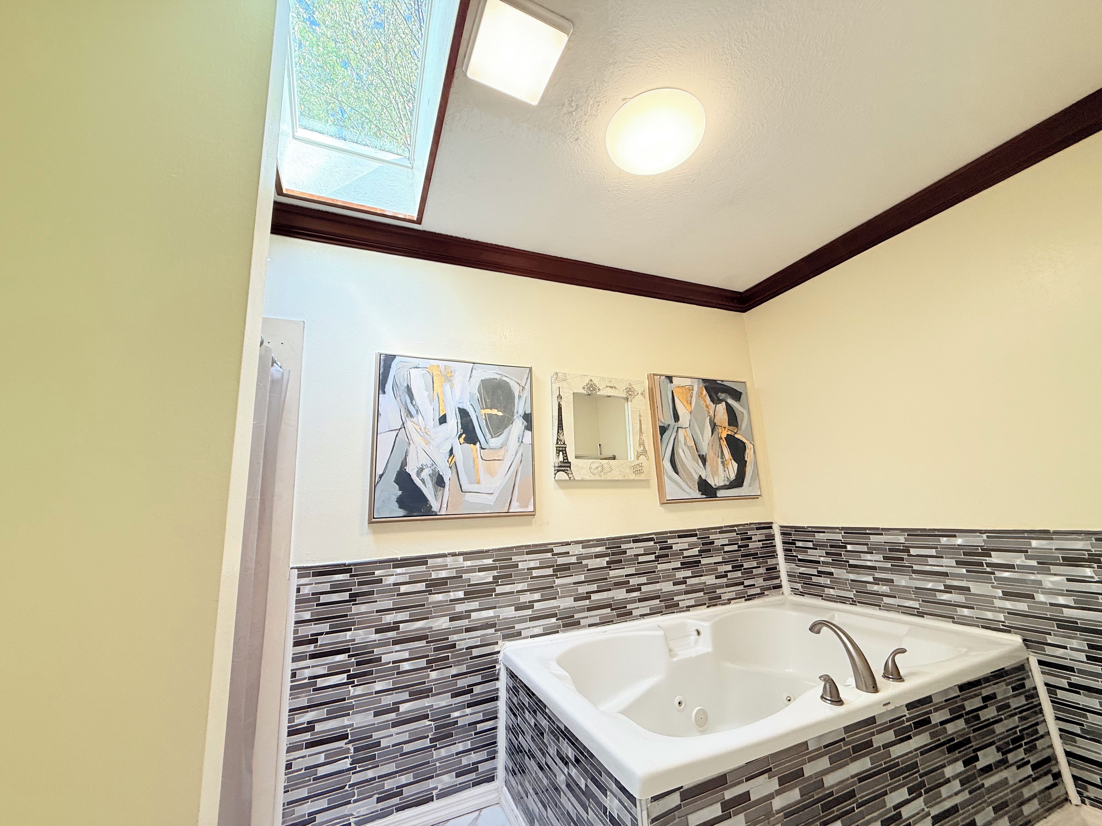 A bathroom with a large bathtub, mosaic tile backsplash, and two abstract paintings on the wall.