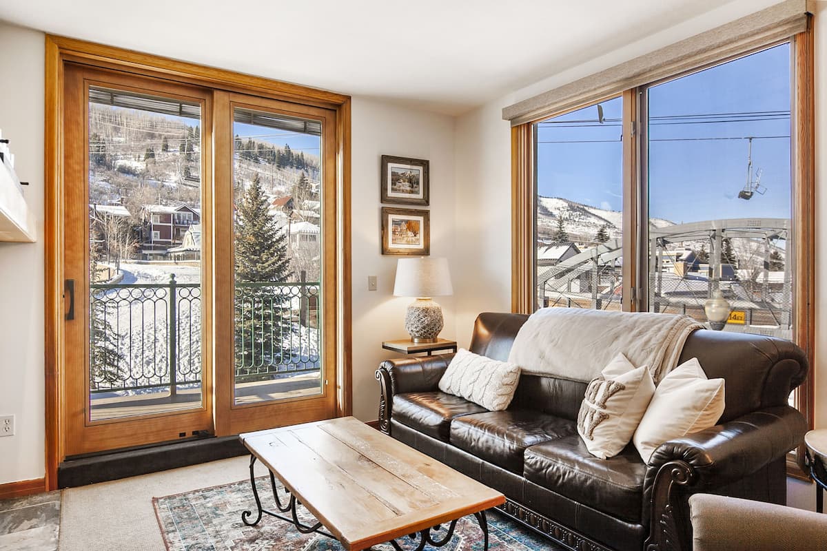 Ein gemütliches Wohnzimmer mit einem Ledersofa, einem Holztisch und Blick auf eine verschneite Berglandschaft durch große Fenster.