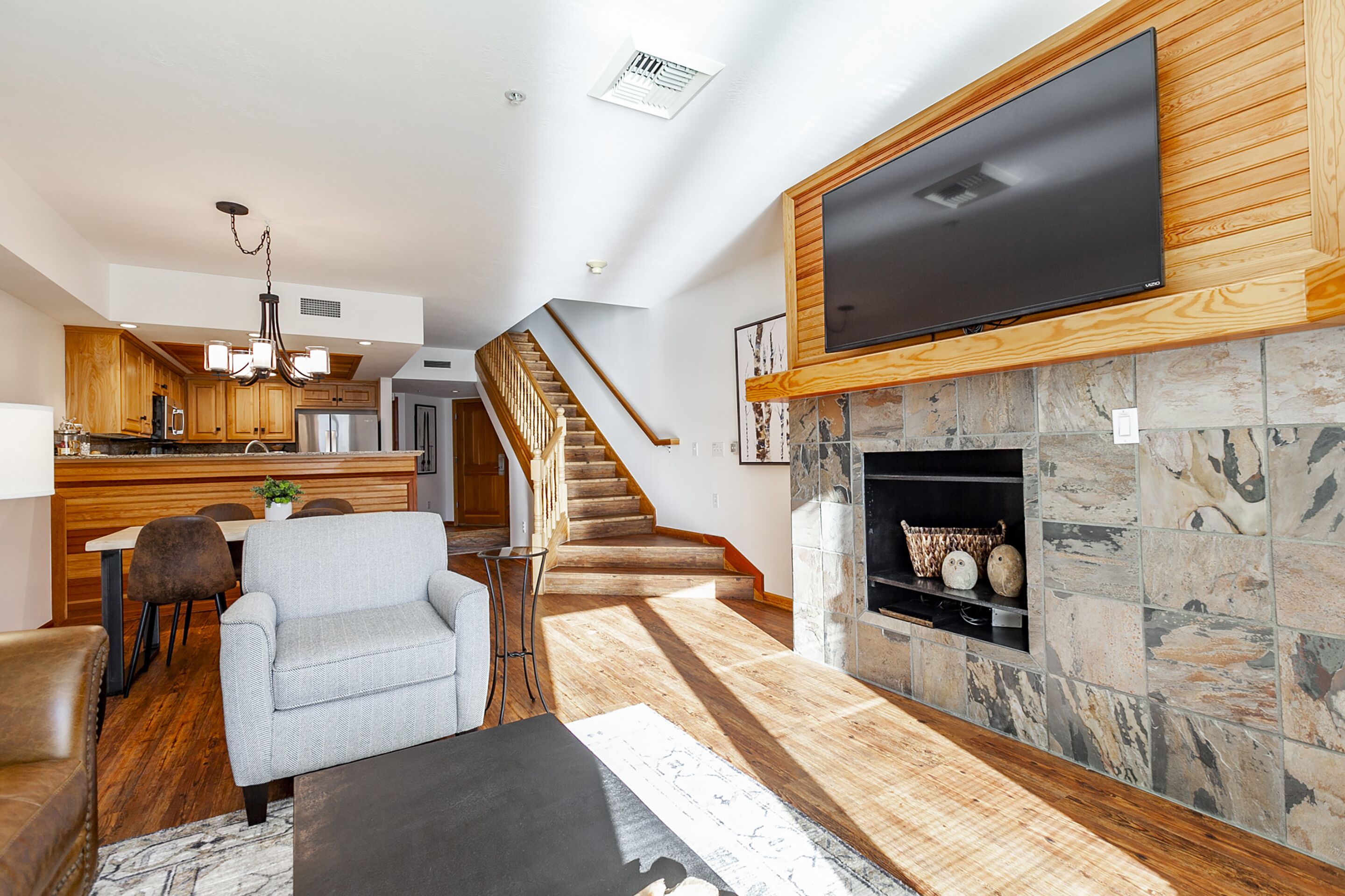 A modern living room with a fireplace, a flat-screen TV mounted on a wooden wall, a staircase leading to an upper level, and a kitchen area with wooden cabinets and a dining table.