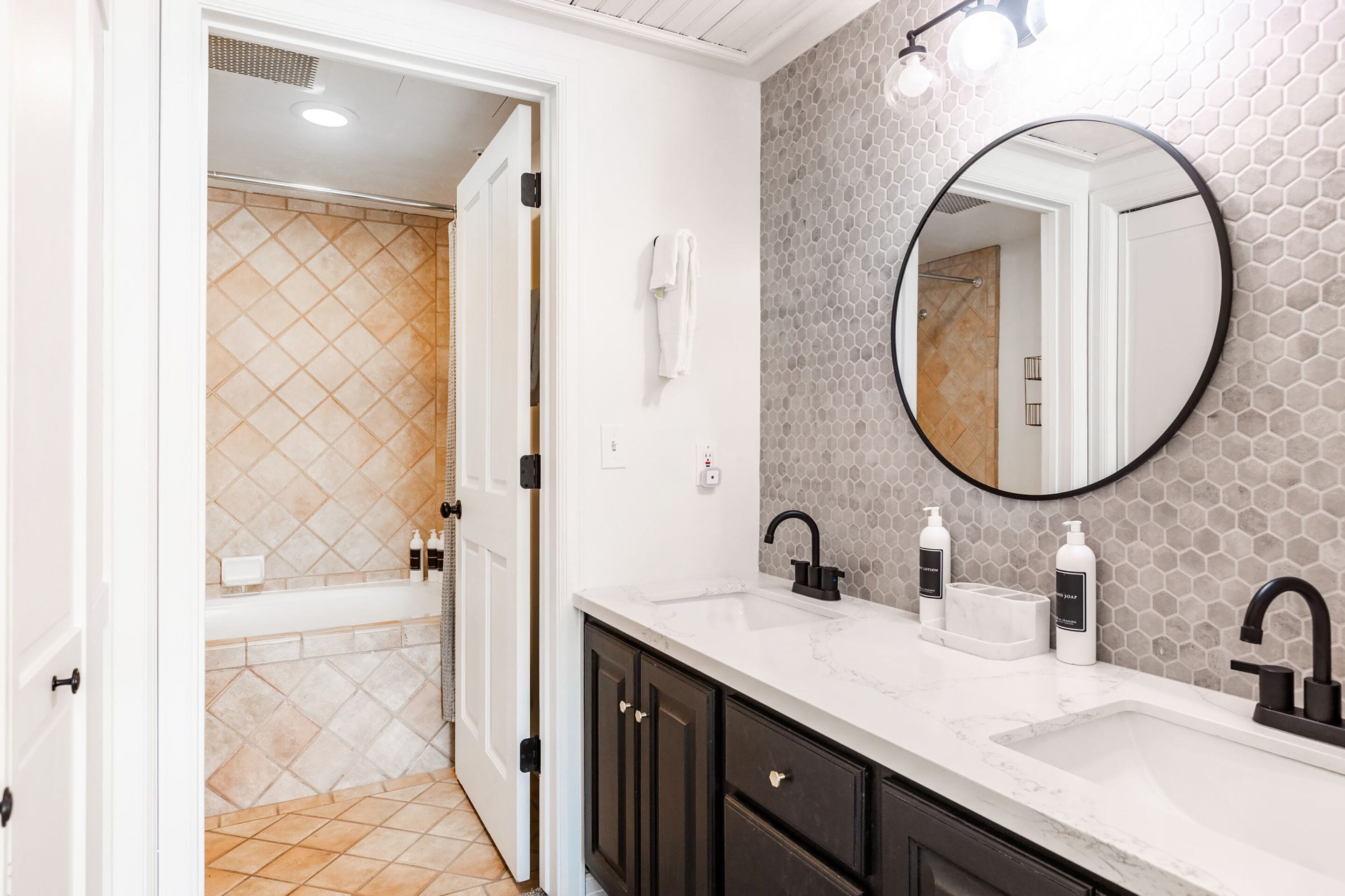 A bathroom with a white vanity, two sinks, a round mirror, and a shower area with beige tiles.
