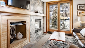 Een moderne woonkamer met een open haard, een flatscreen televisie, een groot raam met uitzicht op een besneeerd landschap, en een gezellige zithoek.