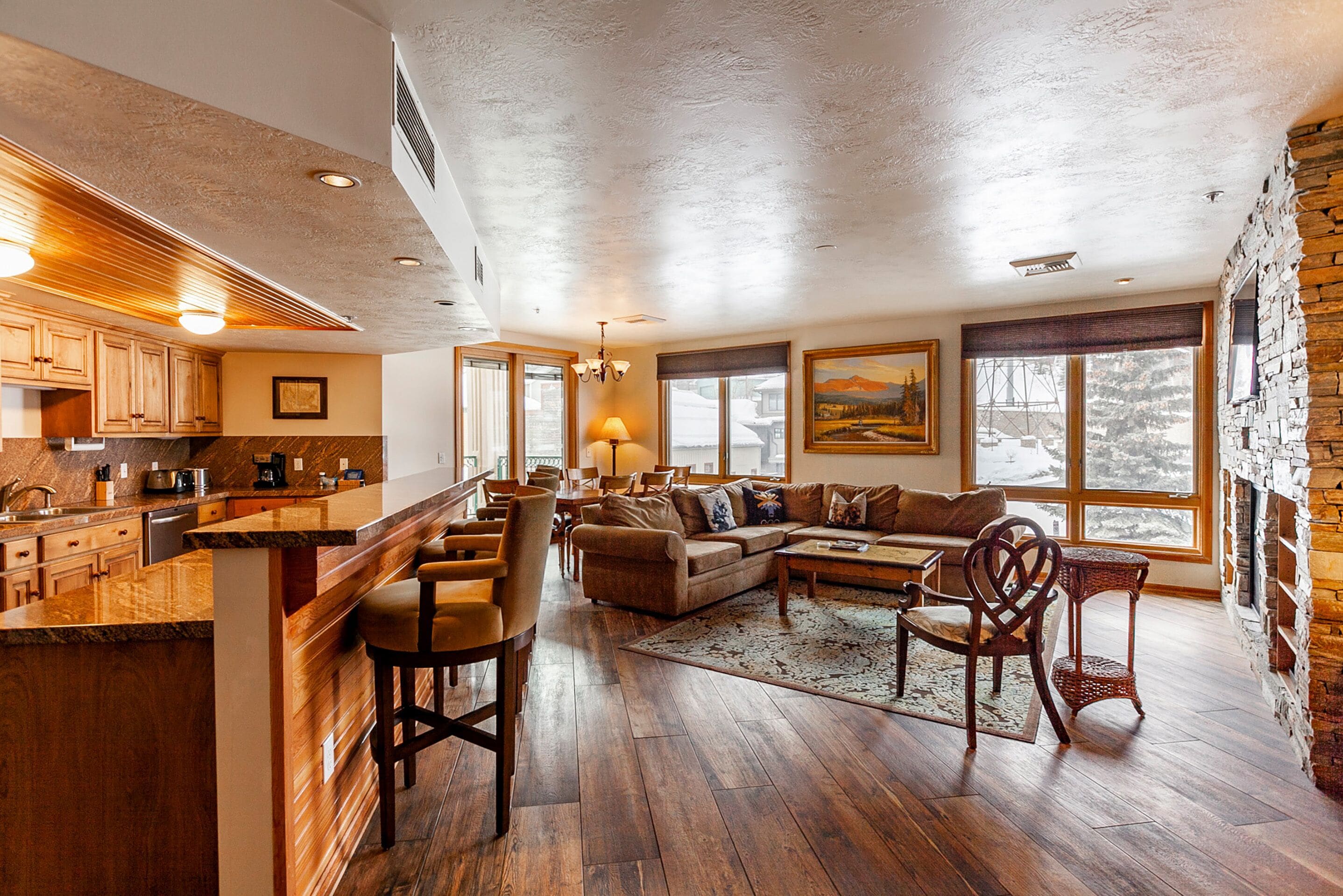 Un espacio habitable amplio con piso de madera, una cocina con gabinetes de madera, una zona de bar y ventanas grandes que ofrecen vistas al exterior.