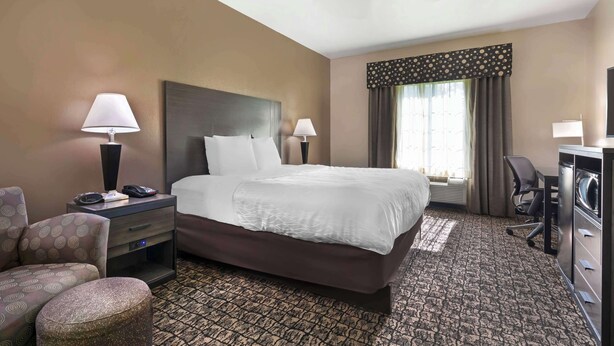 A hotel room with a large bed, two bedside lamps, a desk with a chair, a small round ottoman, and a ceiling fan.