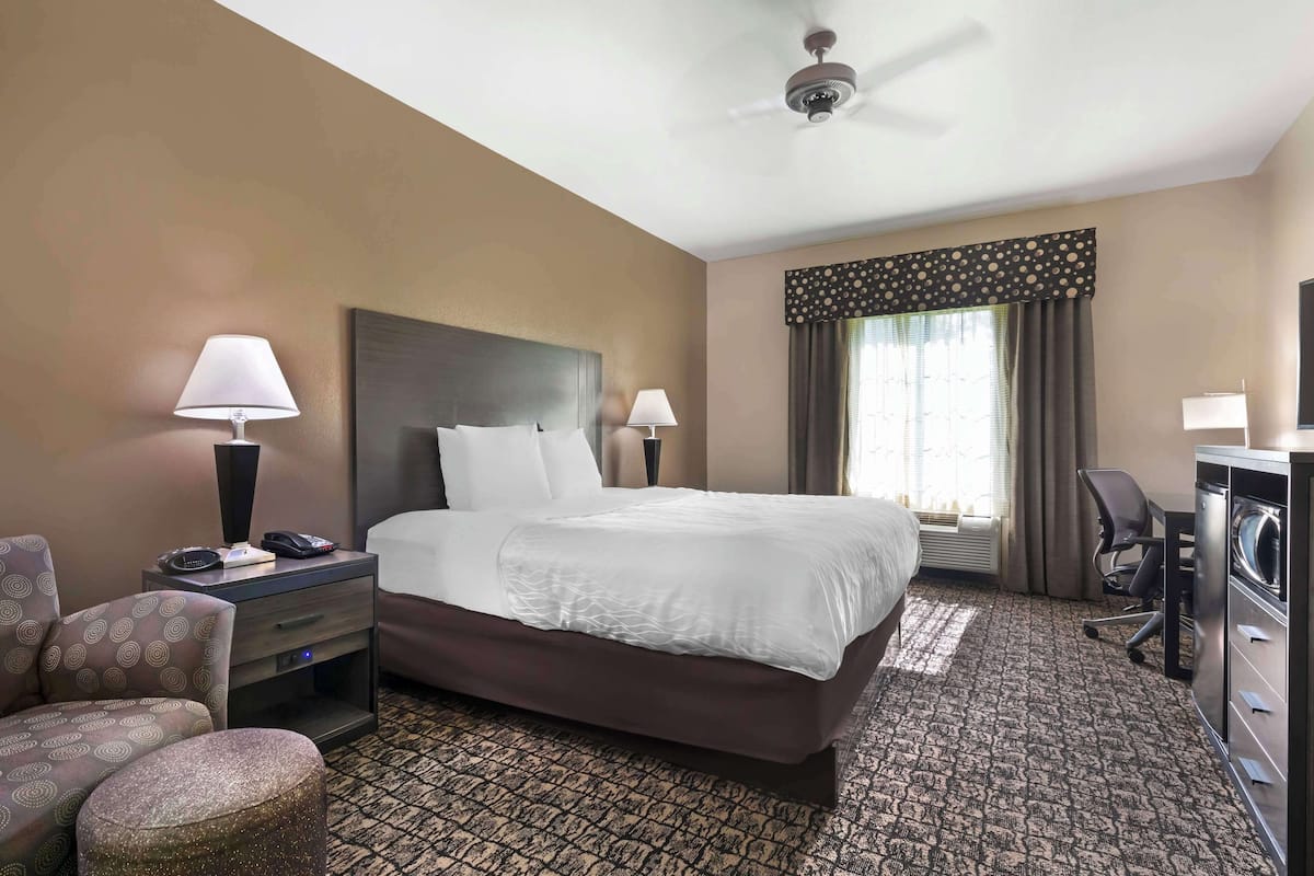 A hotel room with a large bed, two bedside lamps, a desk with a chair, a small round ottoman, and a ceiling fan.