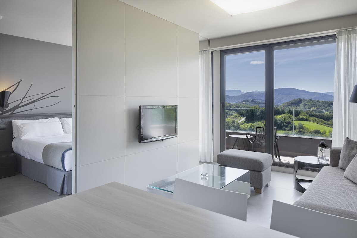 A modern hotel room with a bed, a TV, a glass coffee table, a sofa, and a balcony with a scenic view.