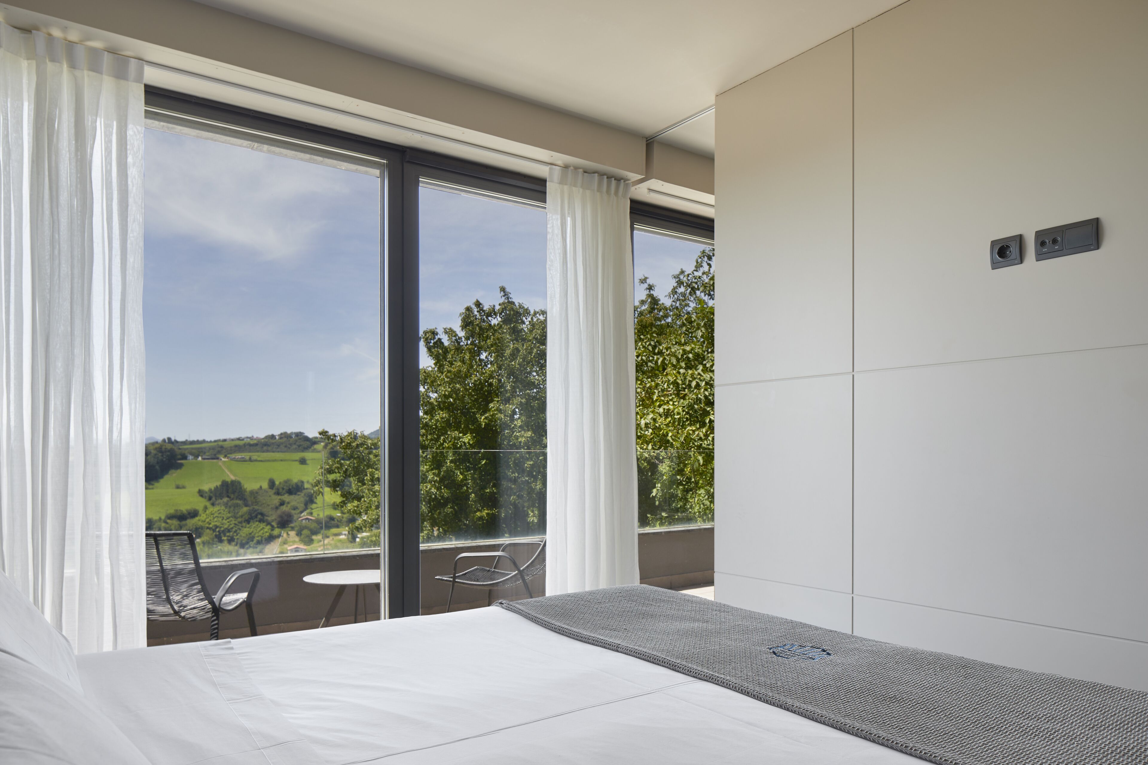 A hotel room with a large bed, a balcony with a view of greenery, and a small table.