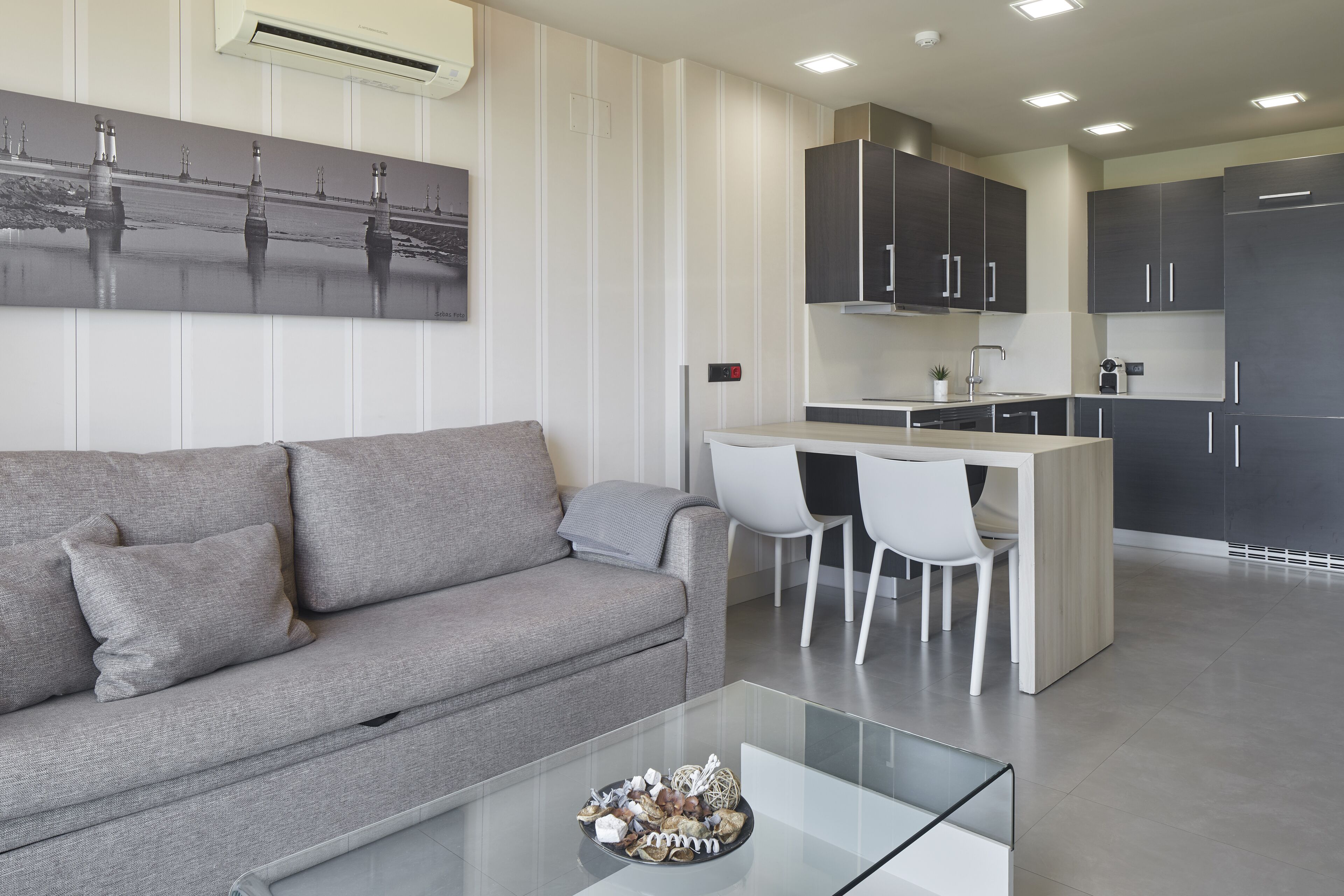 A modern living room with a grey sofa, a glass coffee table, and a kitchen area with dark cabinets and white chairs.