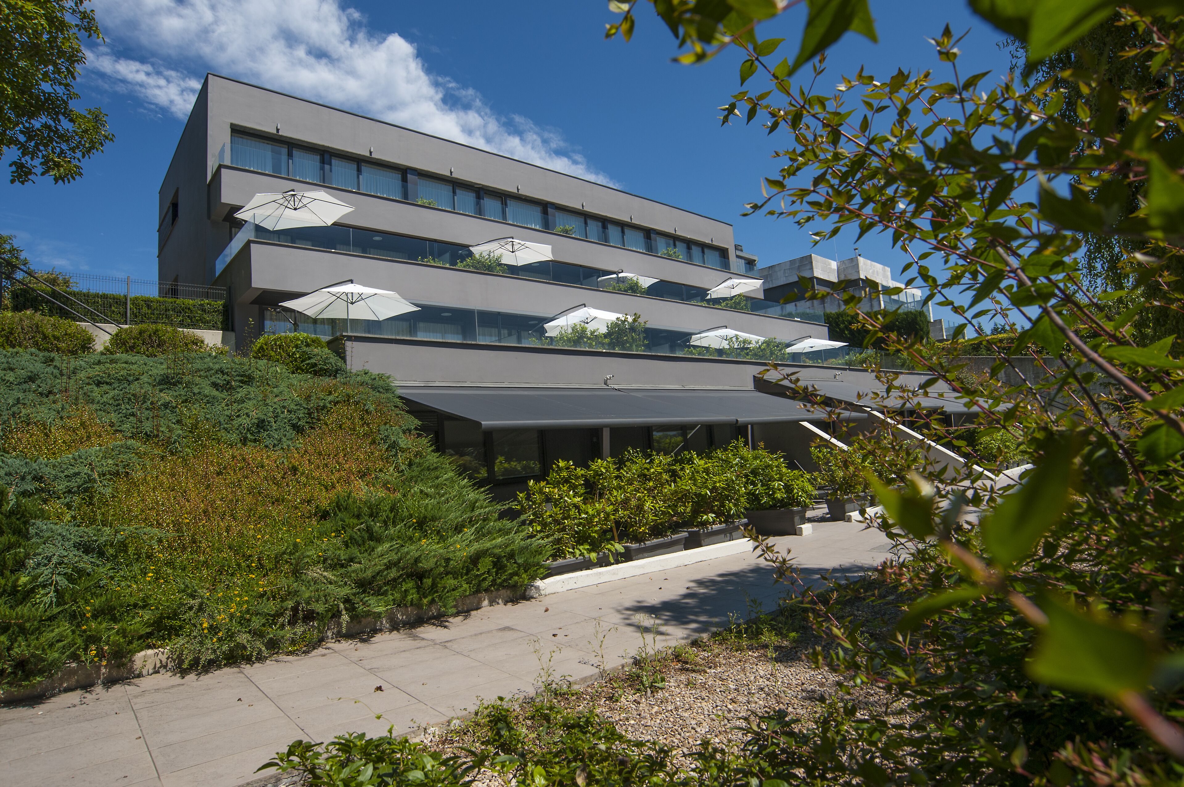 A modern building with multiple balconies, outdoor seating areas with umbrellas, and a landscaped garden.