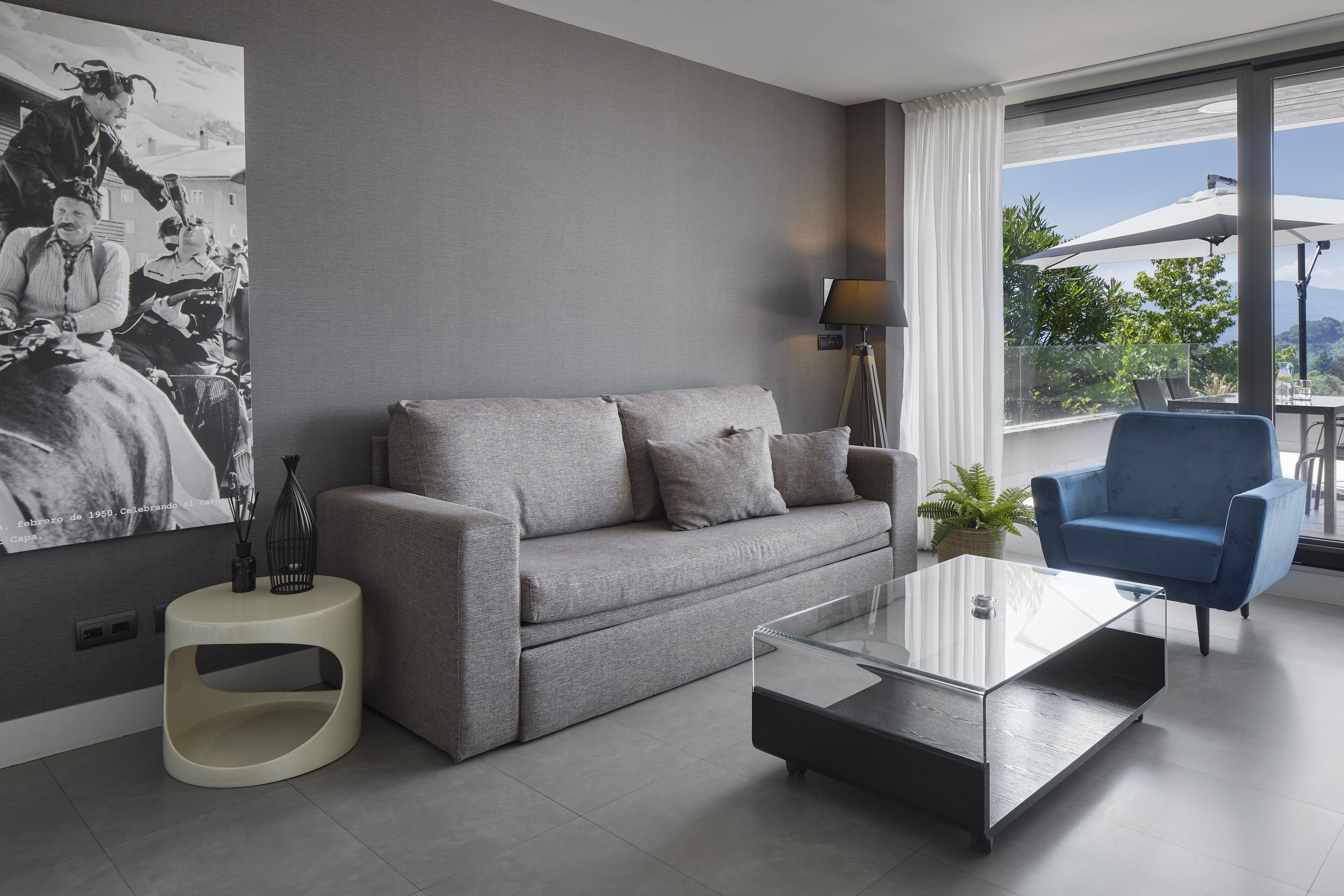 A modern living room with a grey sofa, a glass coffee table, a blue armchair, and a large window with a view of greenery.