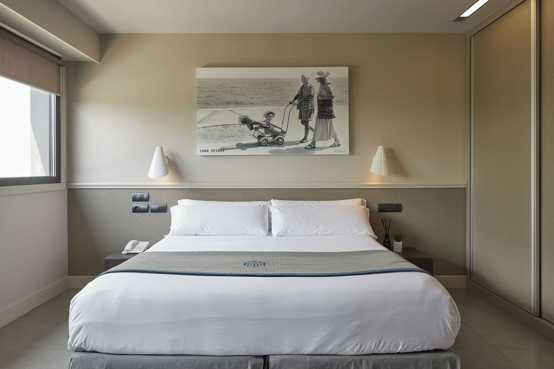 Habitación de hotel con una cama grande, dos lámparas de noche, un cuadro de una playa y una mesita con lámpara.