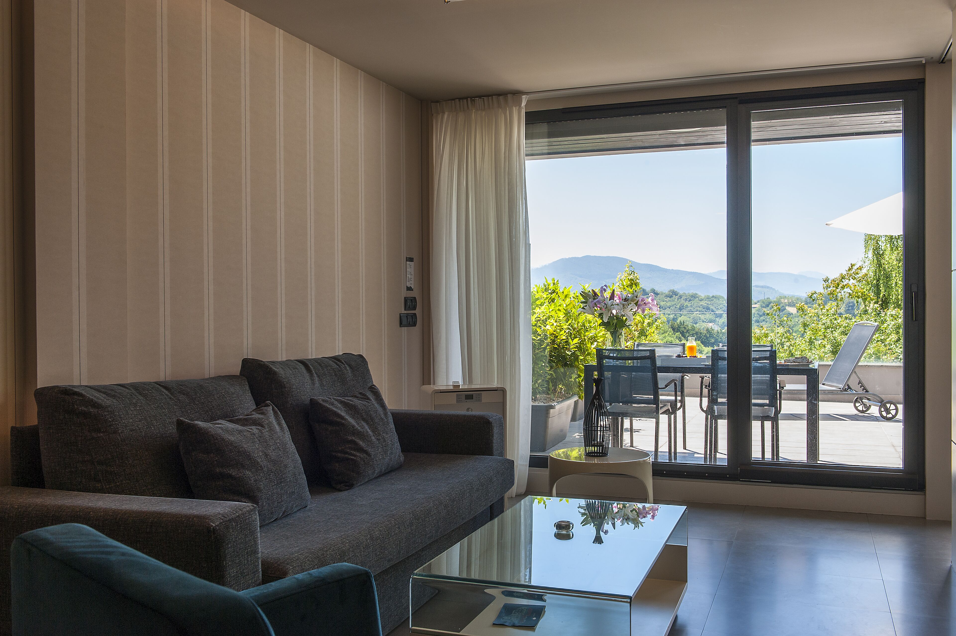 Een moderne woonkamer met een bank, een glazen salontafel en uitzicht op een balkon met buitenmeubilair.