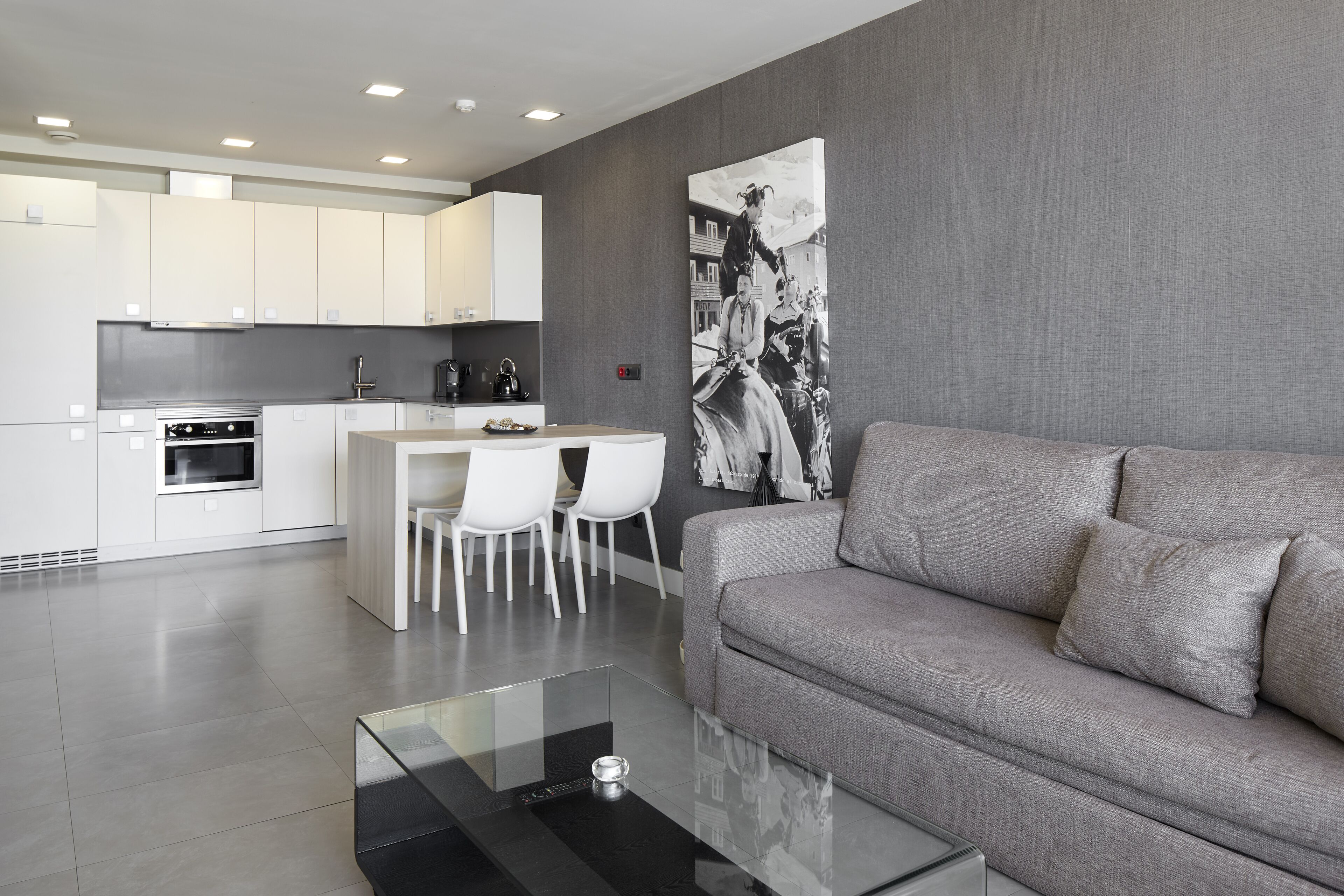 A modern kitchen with white cabinets, a dining area with two white chairs, and a grey sofa in a living area.