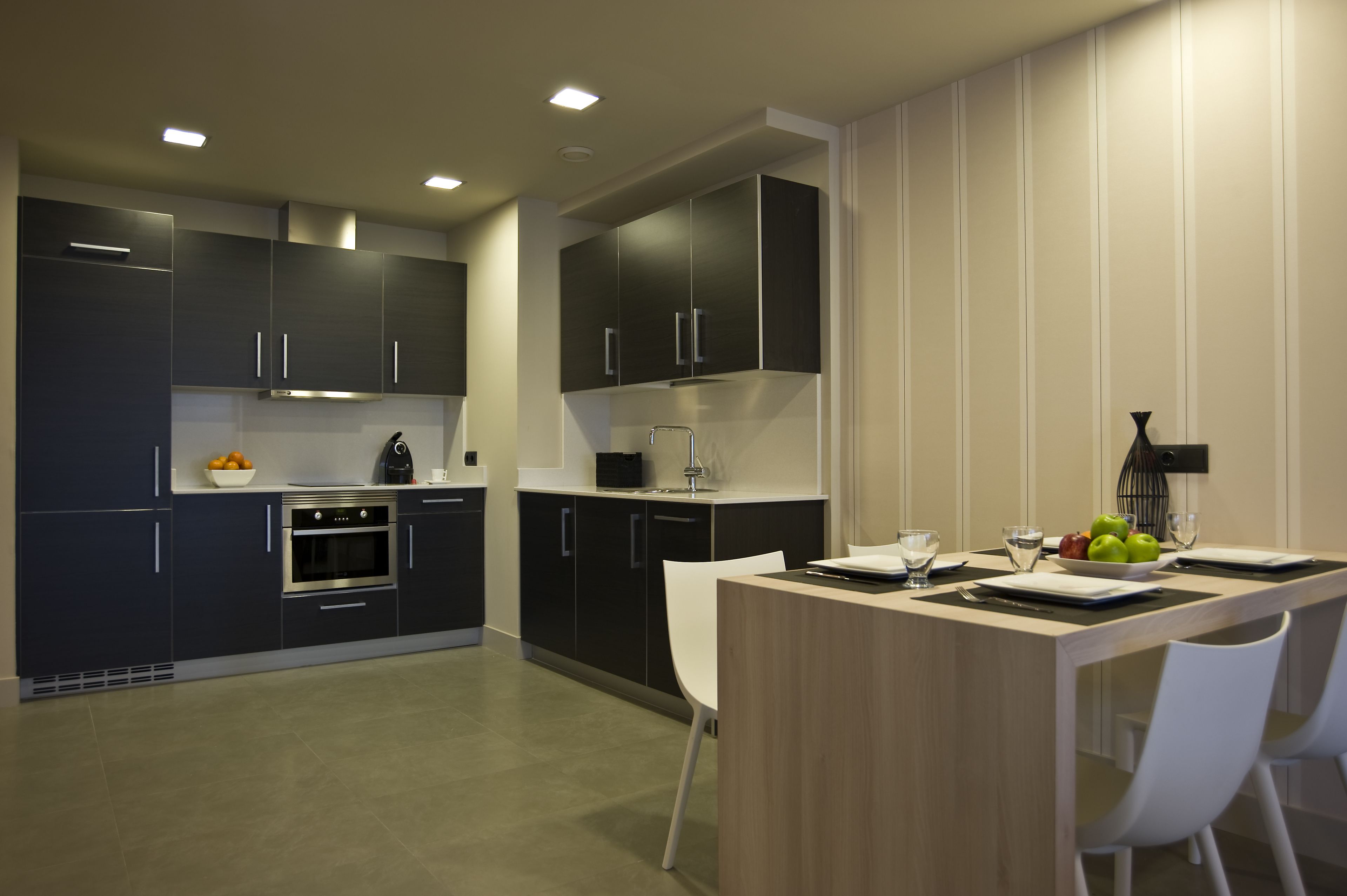 Una cocina moderna con armarios oscuros, horno integrado y comedor con mesa de madera y sillas blancas.