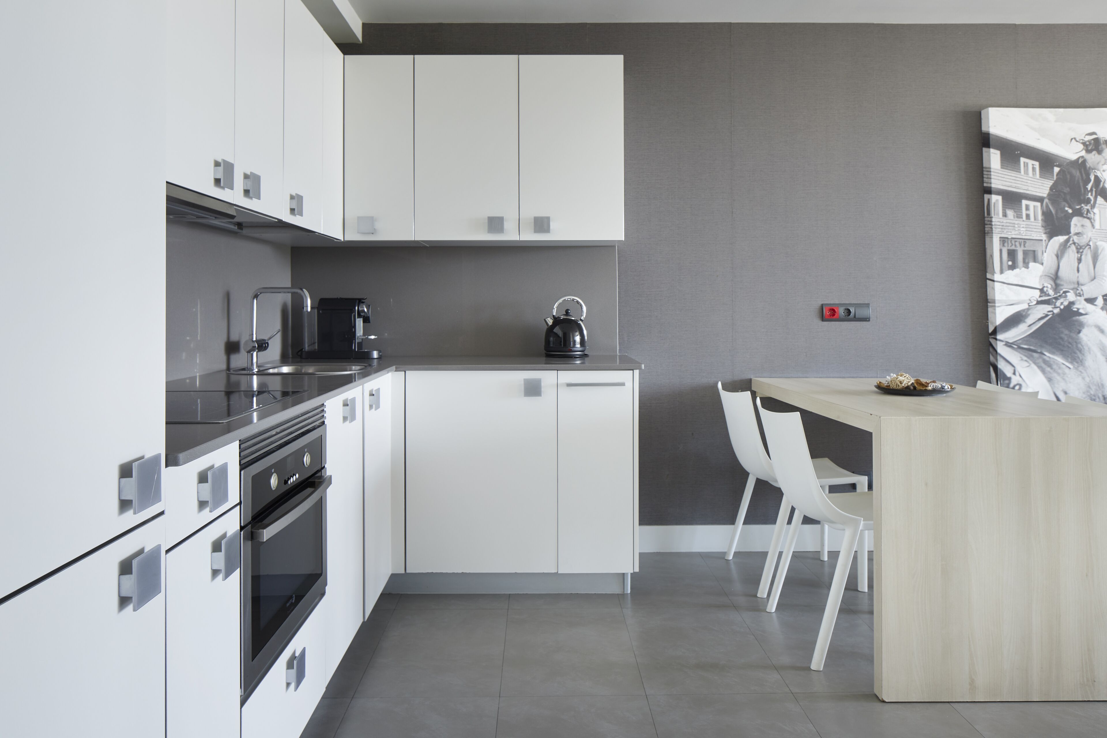 Una cocina moderna con armarios blancos, un hervidor negro y una mesa de comedor de madera con sillas.