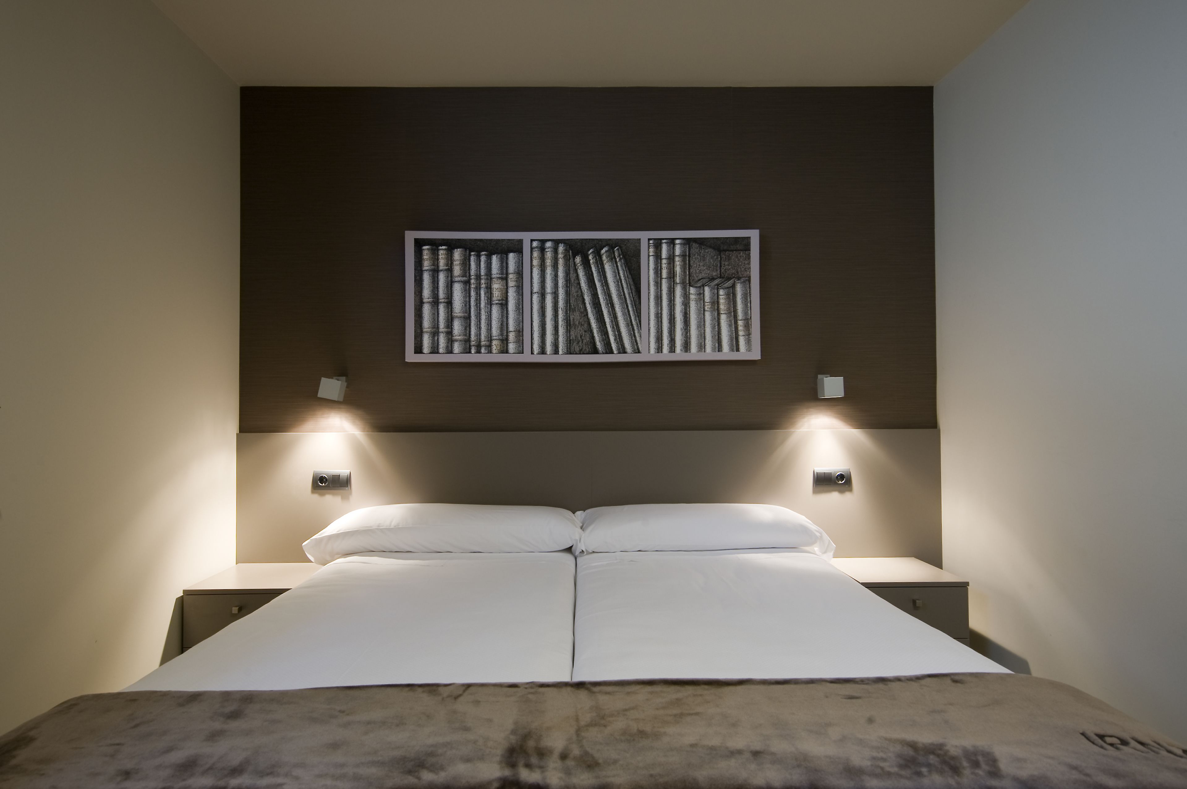 Una cama bien tendida con sábanas blancas, un cabecero con una obra de arte enmarcada y lámparas de noche.