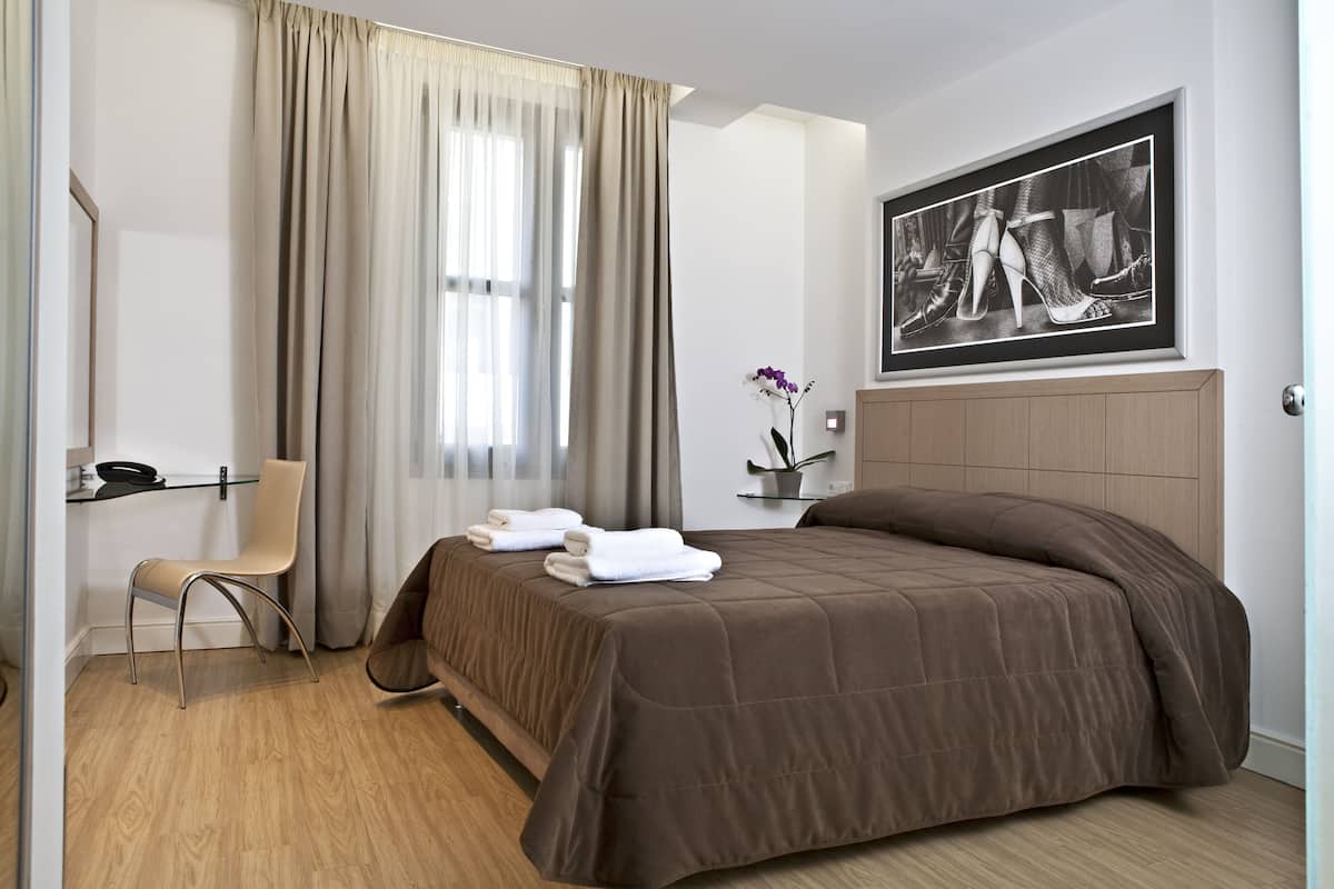 A hotel room with a bed, a chair, a desk, and a window with curtains.